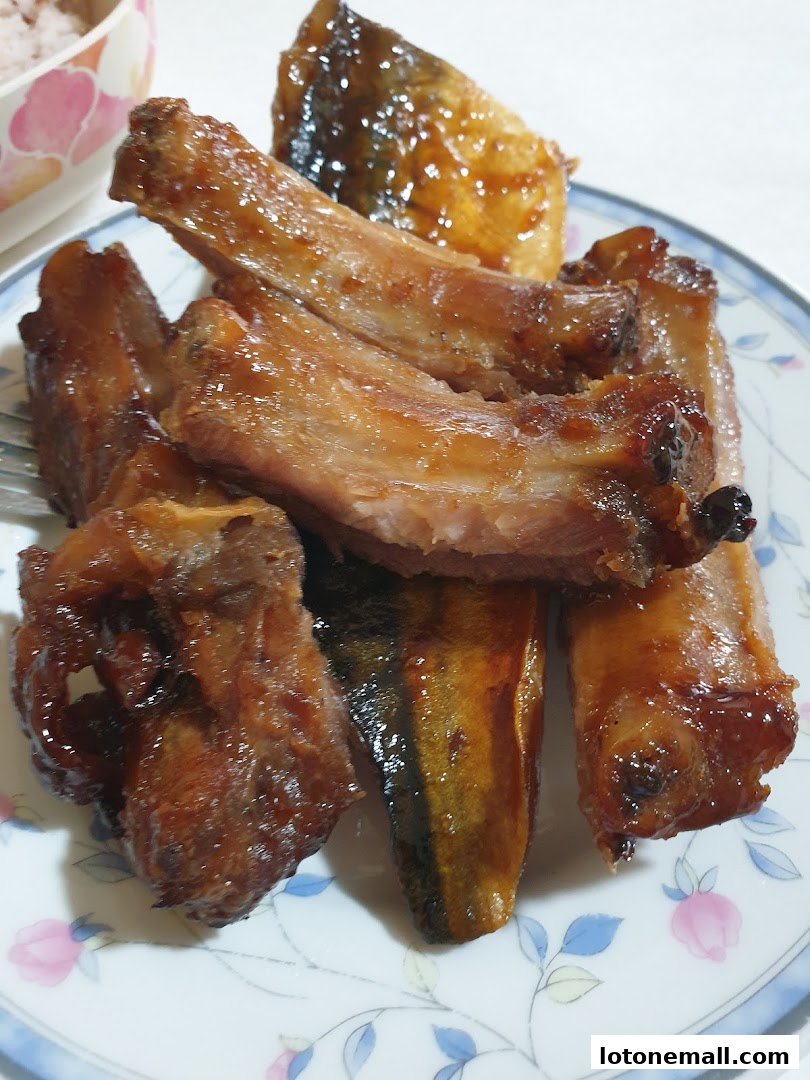 A plate of glazed ribs and fish, with a bowl of rice in the background.