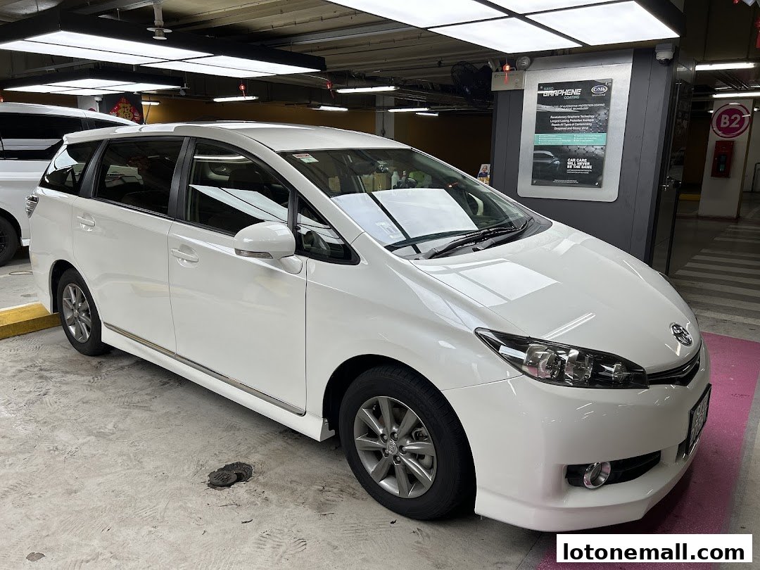 A white Toyota Wish car parked in a parking lot with a promotional poster in the background.