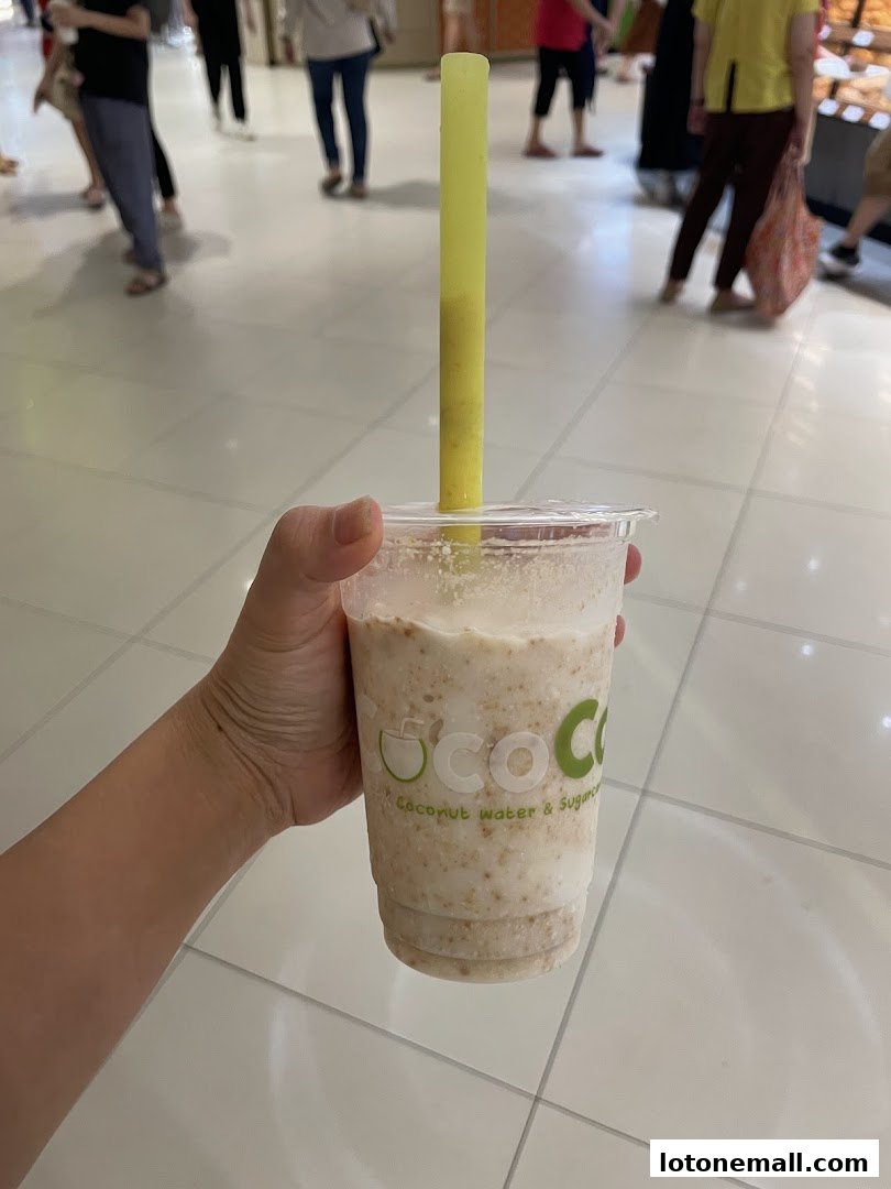 A hand holds a refreshing CocoCane drink with a yellow straw in a mall setting.
