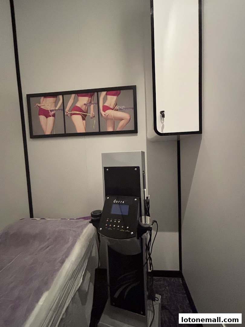 A treatment room with a slimming machine, bed, and before-and-after body measurement posters.
