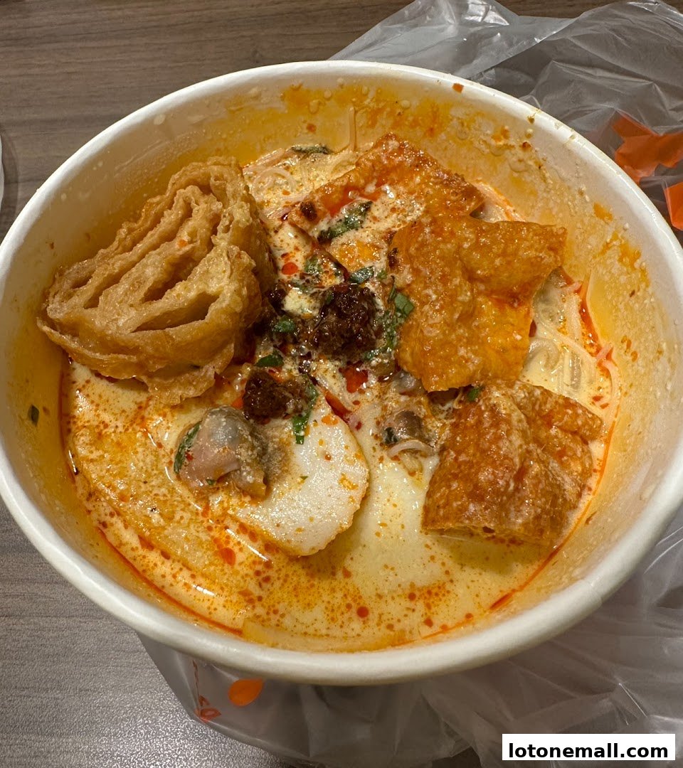 A close-up of a bowl of Laksa with fish cakes, fried tofu puffs, and chili oil.