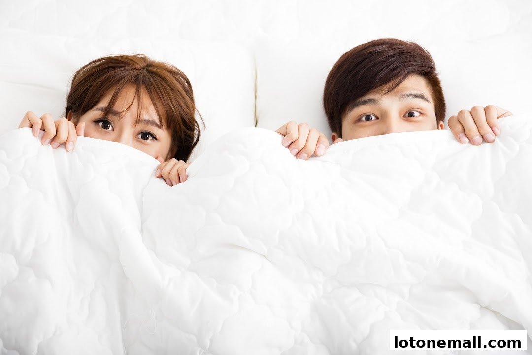 A couple peeking out from under a white duvet, looking surprised.