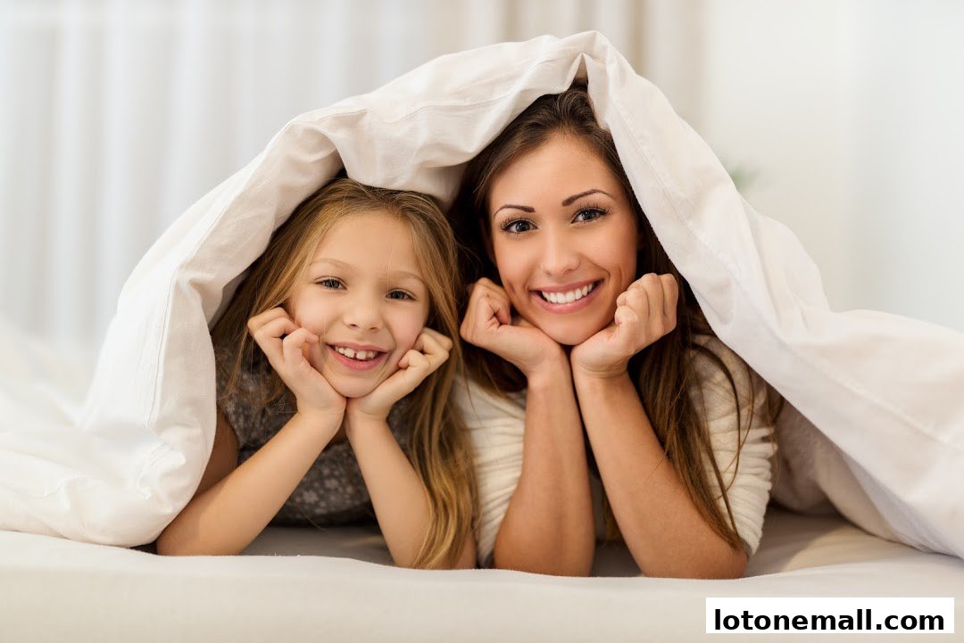 A mother and daughter smile happily from under a white duvet.