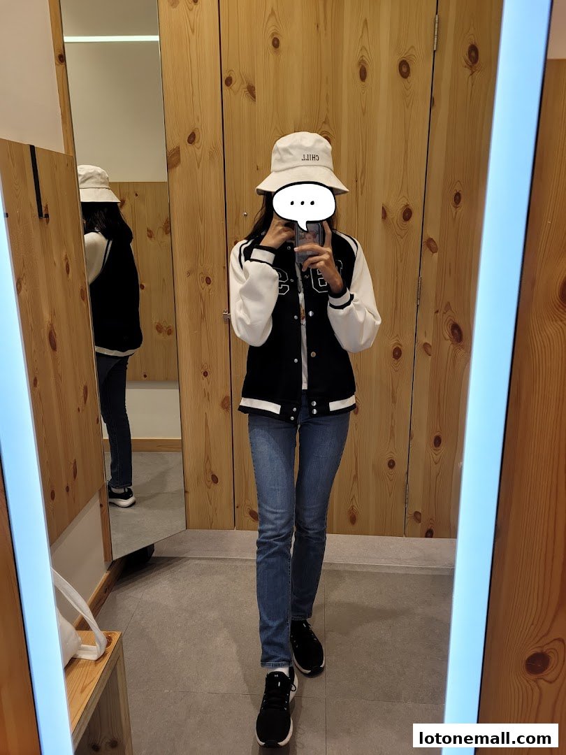 A person in a fitting room wearing a varsity jacket, jeans, and a bucket hat.