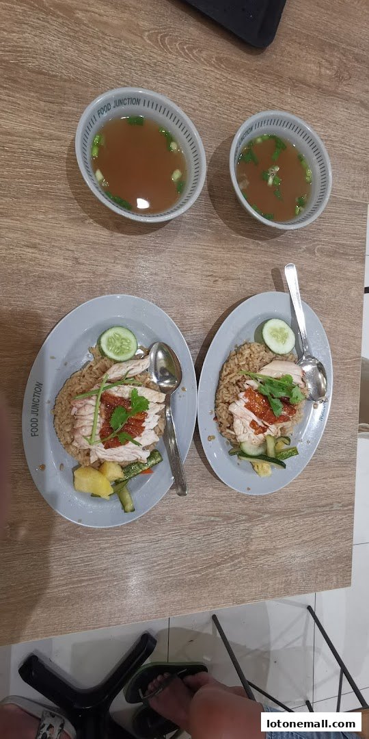 Two plates of chicken rice with soup and cucumber slices on a wooden table.