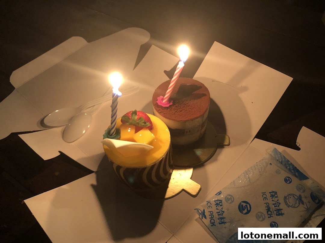 Two birthday cakes with lit candles, spoons, and an ice pack in a box.