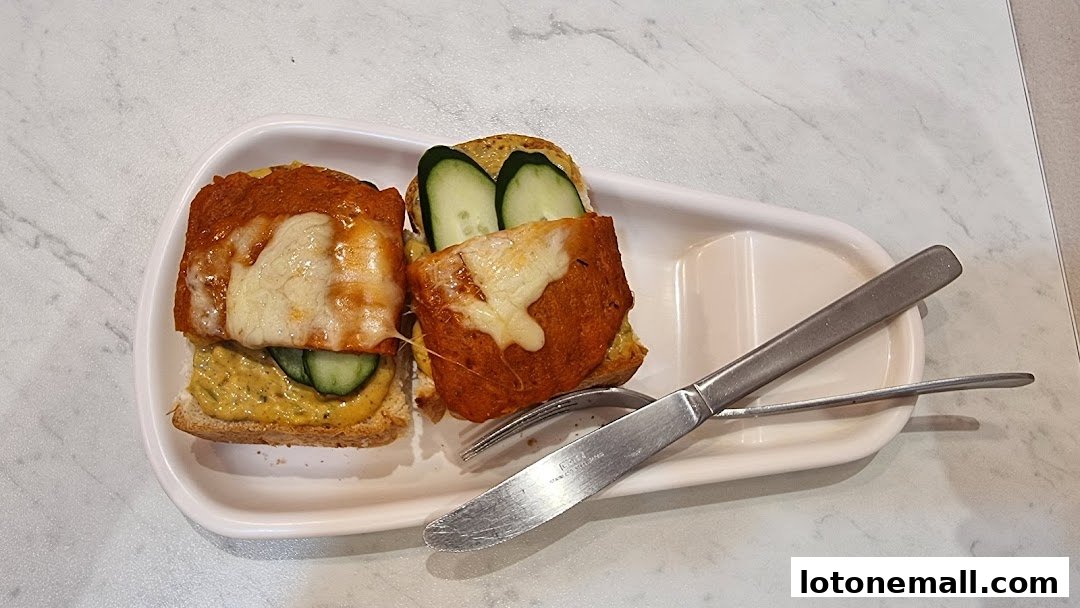 Two open-faced sandwiches with cheese, sauce, and cucumber on a white plate with cutlery.