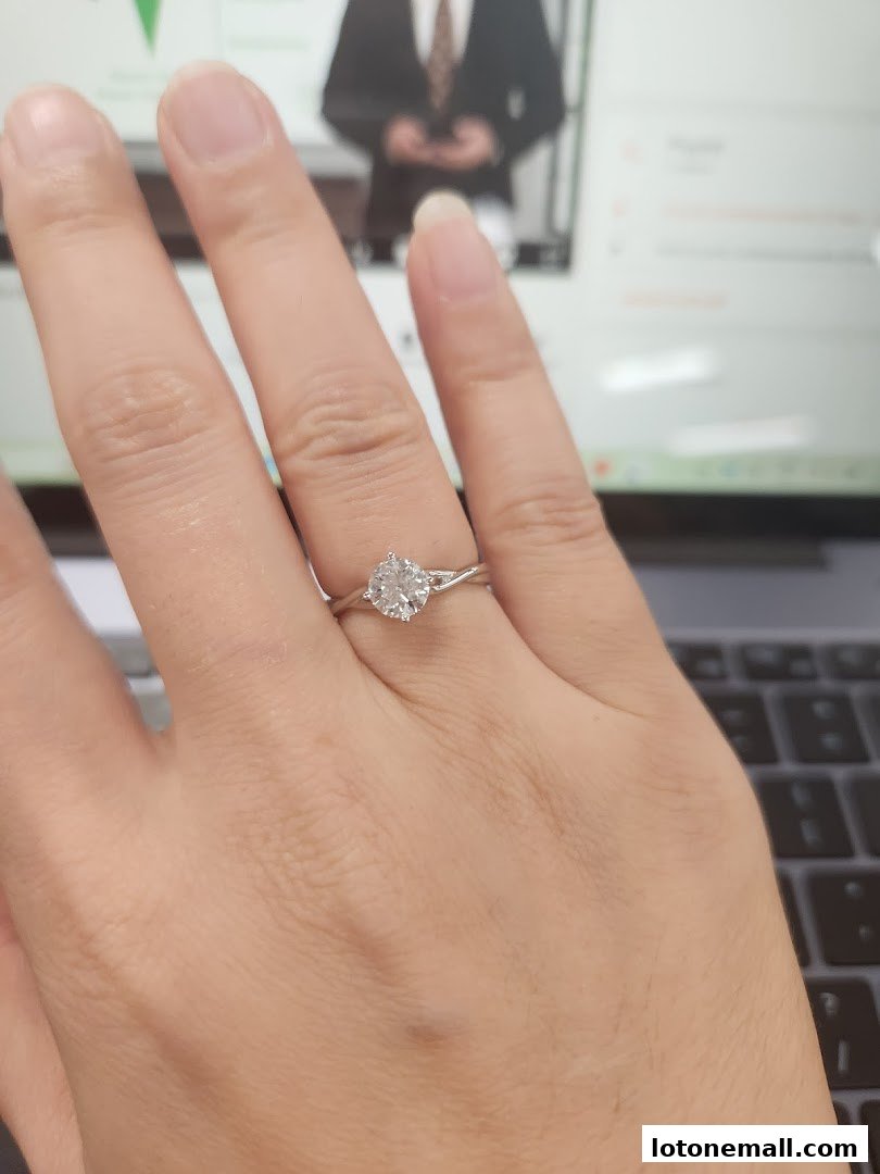 A hand wearing a diamond engagement ring with a laptop in the background.