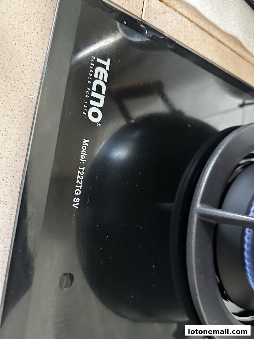 Close-up of a black Tecno gas stove with a blue flame and model number visible.