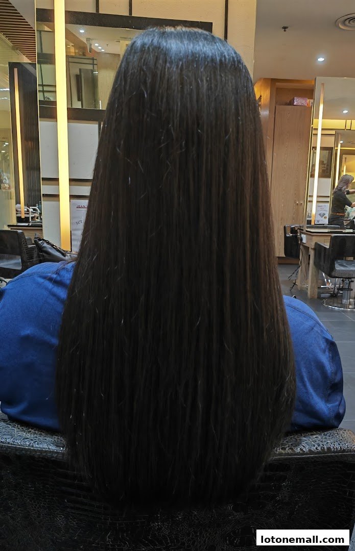 A person with long, dark, straight hair sitting in a salon chair, viewed from behind.