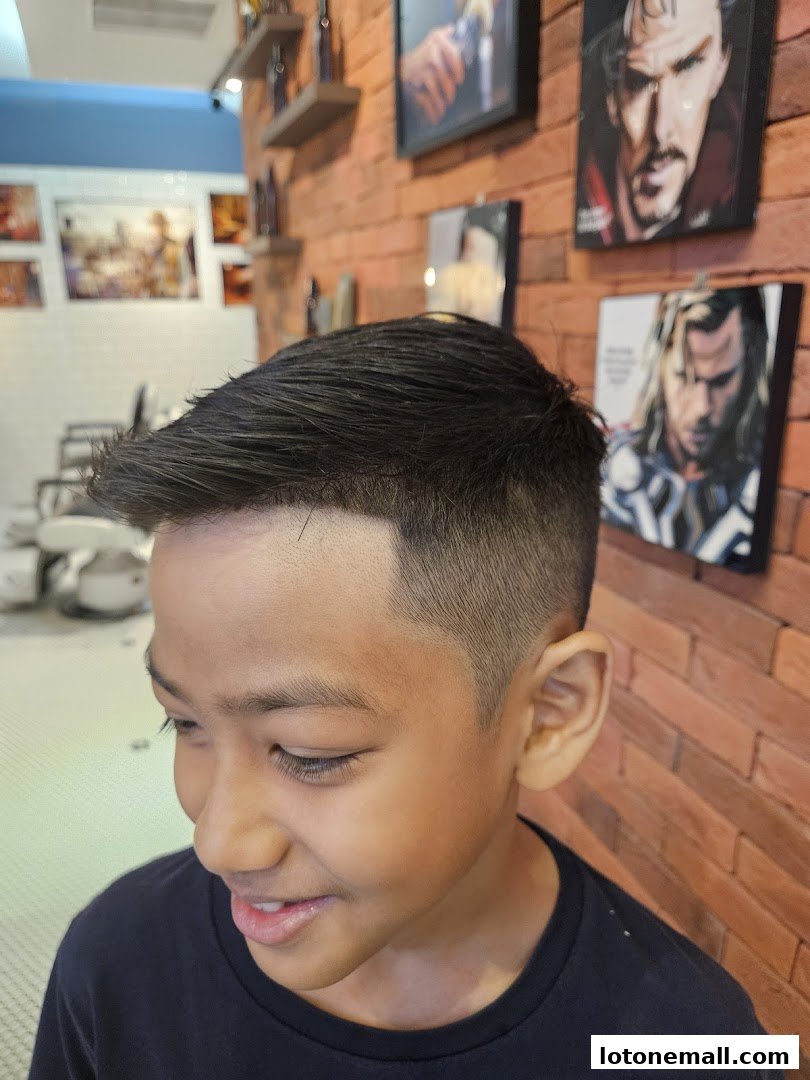 A young boy with a fresh haircut smiles in a barbershop with superhero art on the wall.