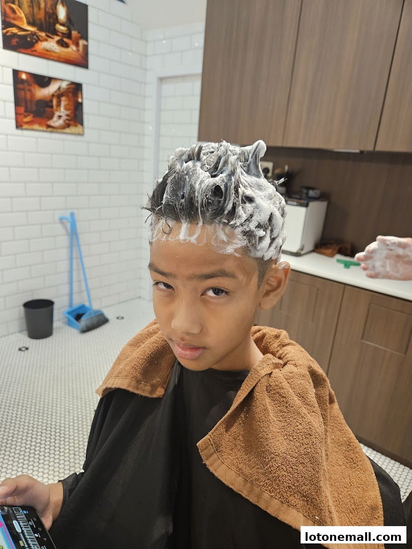 A young boy with shampoo in his hair sits in a barber chair, looking at his phone.