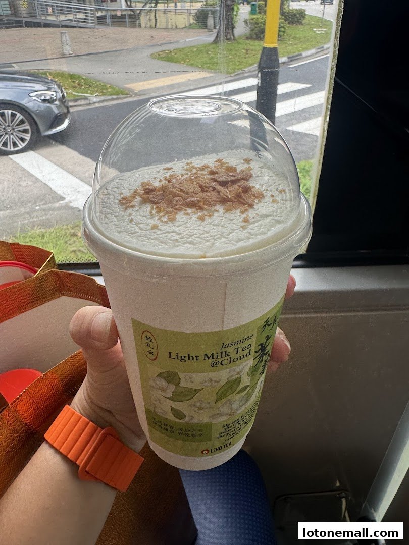 A hand holds a LiHO TEA cup with Jasmine Light Milk Tea @Cloud topped with crunchy bits.