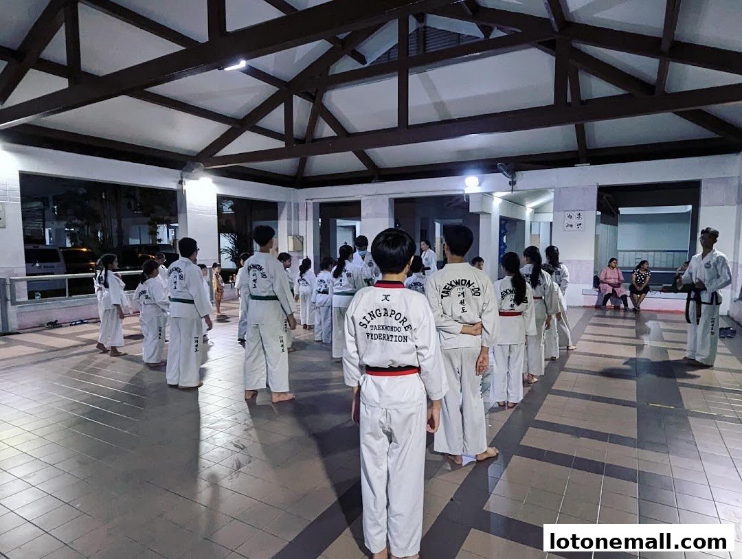A group of children and adults in Taekwondo uniforms stand in a hall, listening to an instructor.