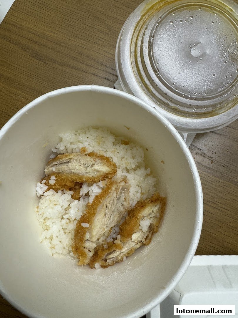 A bowl of rice topped with crispy fried chicken pieces, with a takeout container lid in the background.