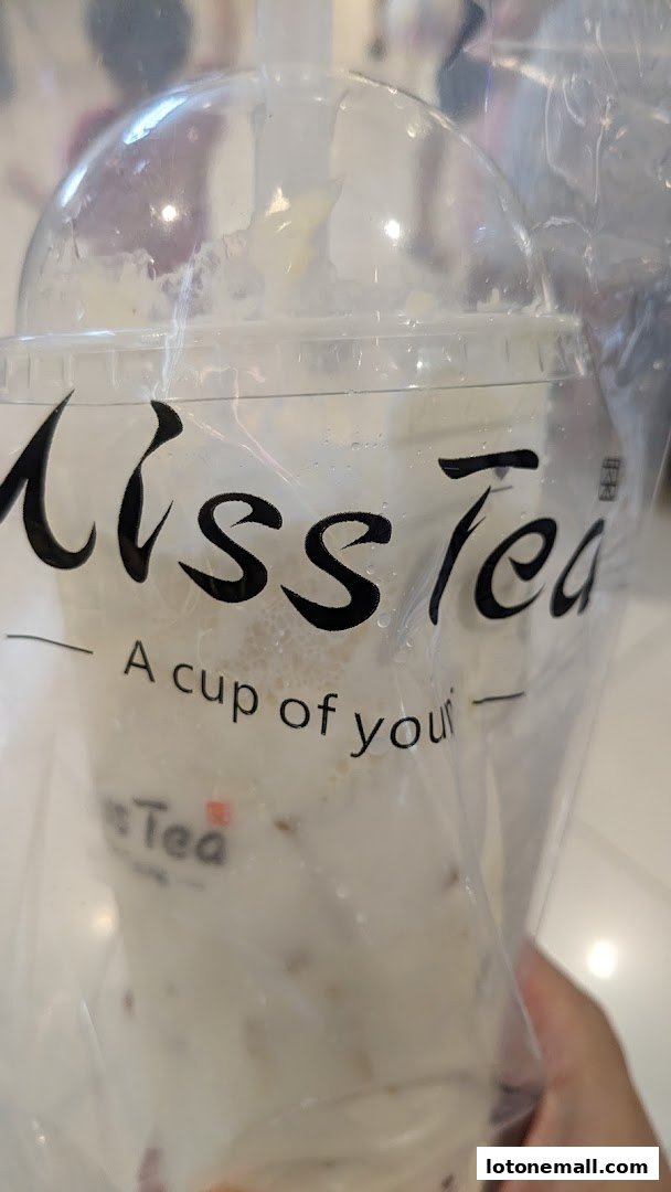 A hand holds a clear plastic cup with the Miss Tea logo and a creamy beverage inside.