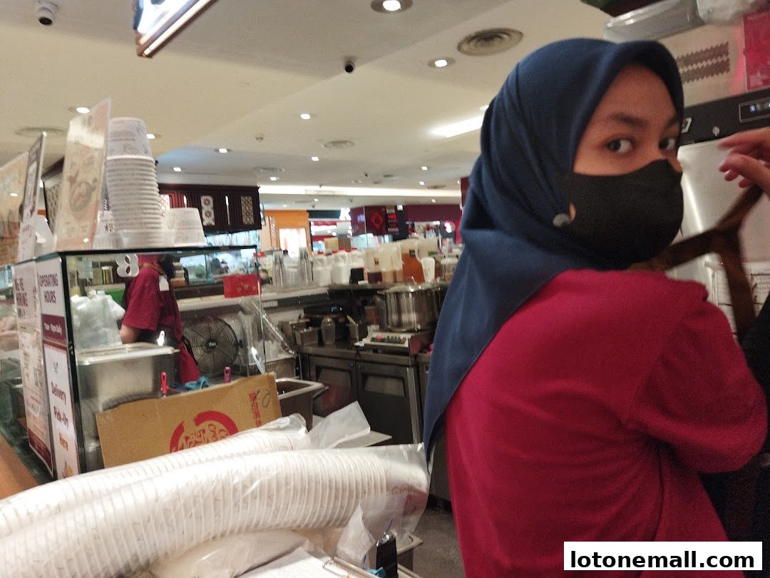 A person wearing a hijab and mask stands in front of a dessert shop counter.