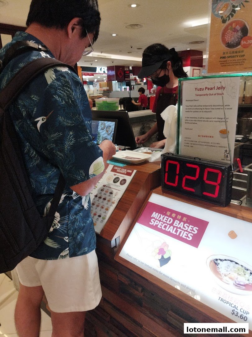 A customer orders at a dessert counter with a menu and a digital queue number display.