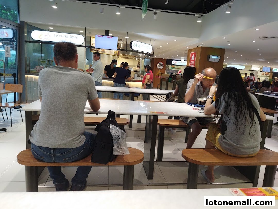 People dining at tables in a food court, with various food stalls visible in the background.