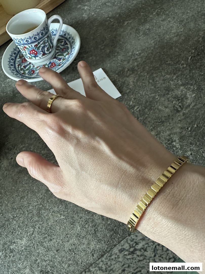 A hand wearing a gold ring and bracelet, with a decorative teacup and saucer in the background.