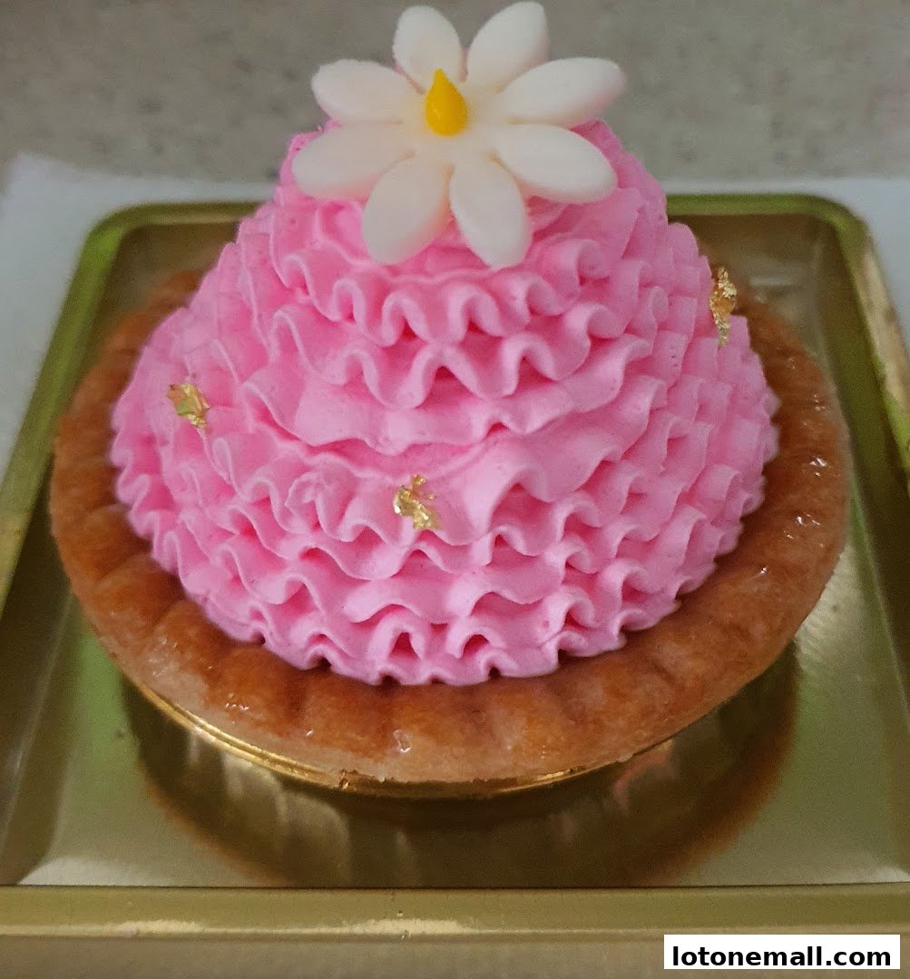 A close-up of a pink frosted tart topped with a white flower and gold leaf.