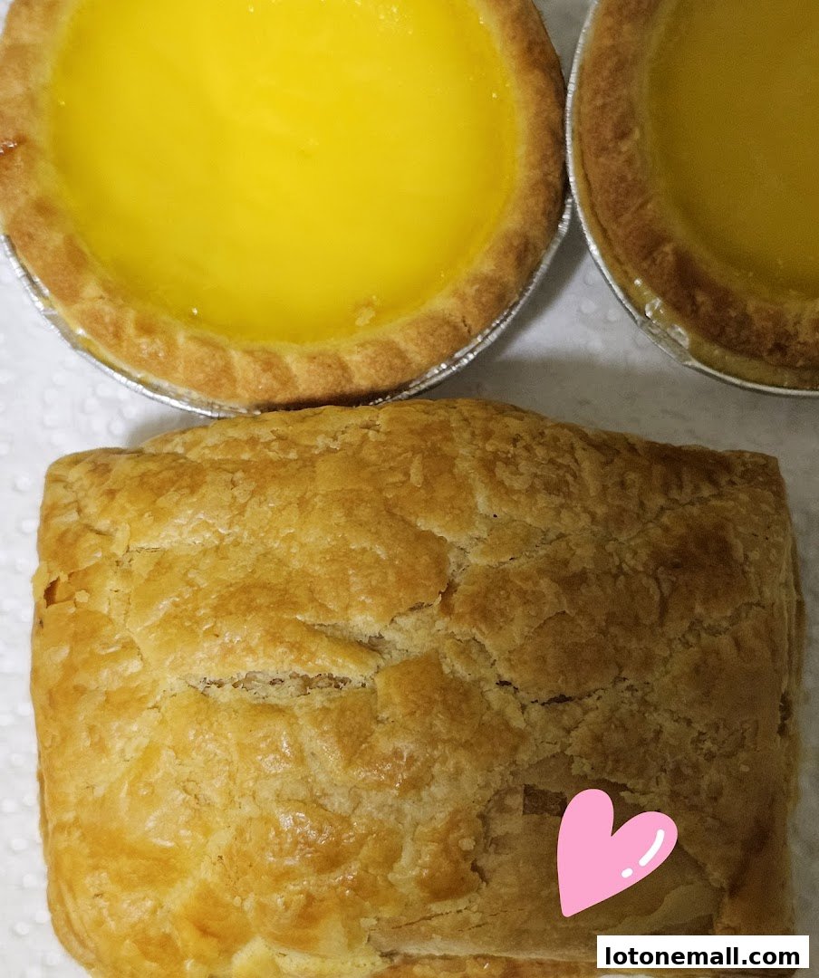 Close-up of two golden egg tarts and a flaky pastry with a pink heart sticker.