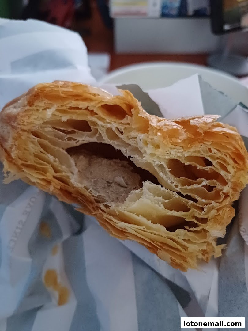 A close-up of a flaky pastry with a filling, wrapped in branded paper.