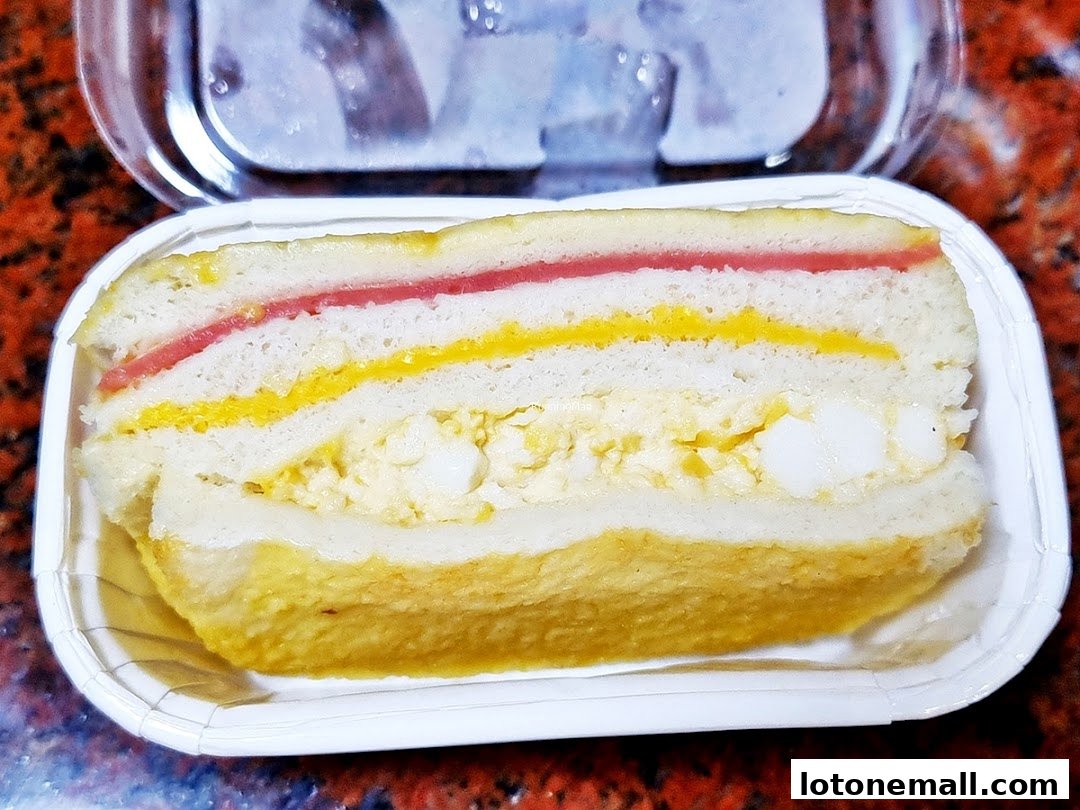 A close-up of a layered sandwich with ham, egg salad, and yellow spread in a white container.