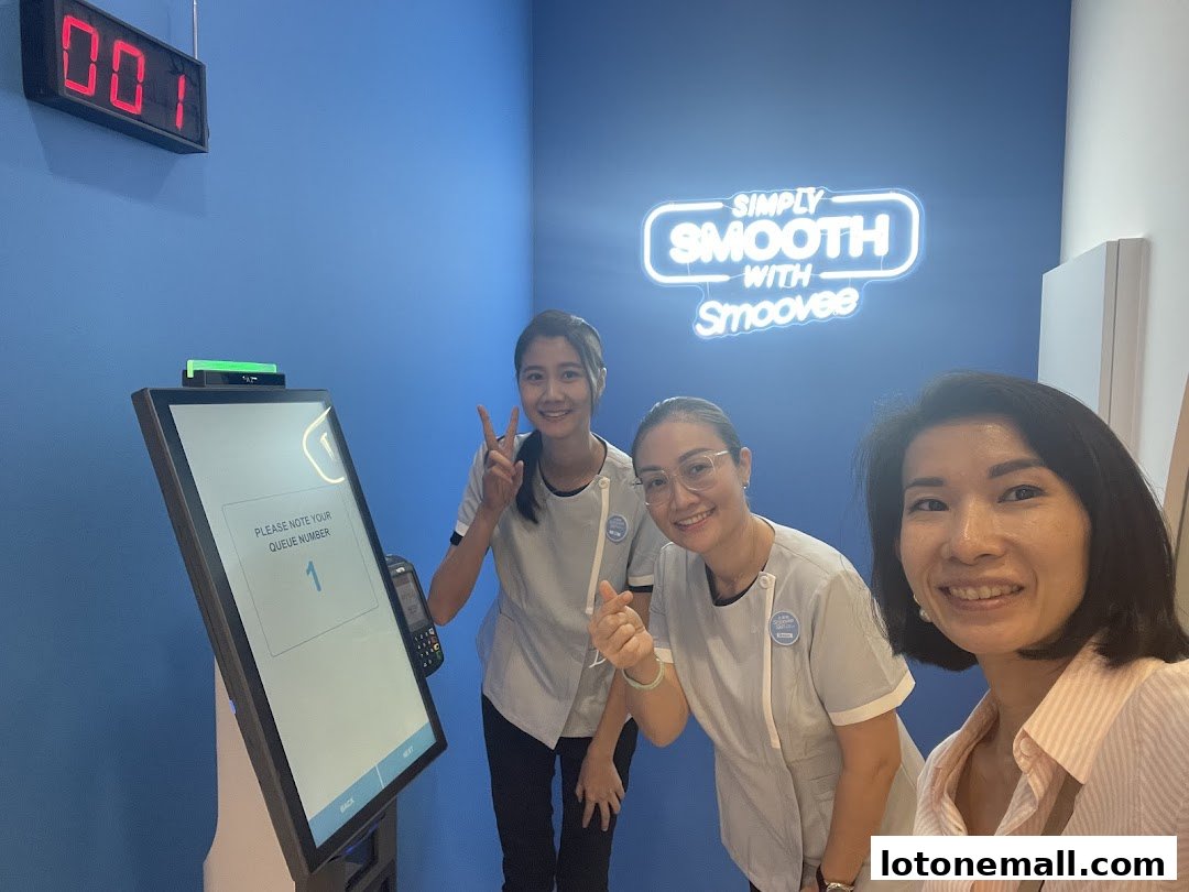 Three smiling women pose for a selfie in front of a queue number display and a neon sign.
