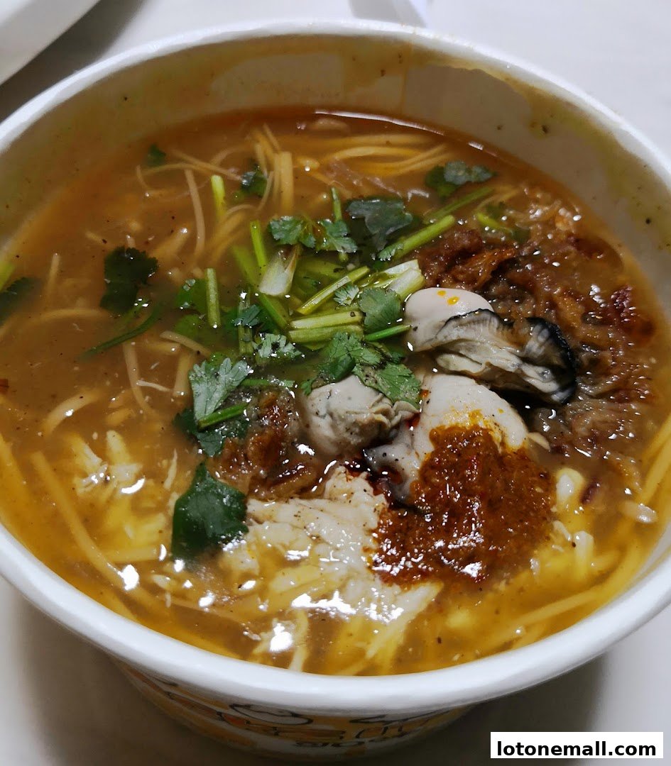 A close-up shot of a steaming bowl of oyster noodle soup with fresh cilantro and chili paste.