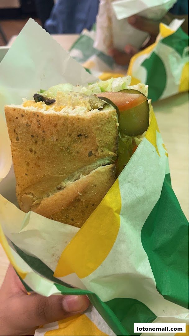 A hand holds a freshly made Subway sandwich wrapped in branded paper.