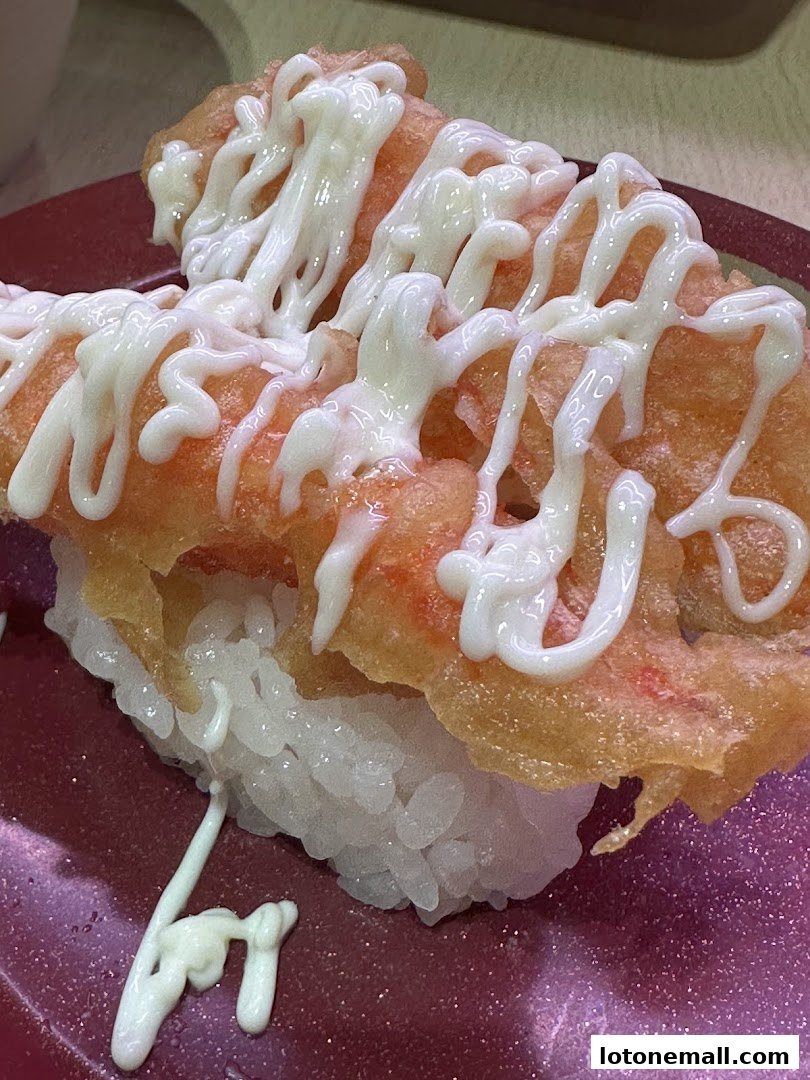 A close-up of a sushi dish with rice, topped with fried ingredients and drizzled with mayonnaise.