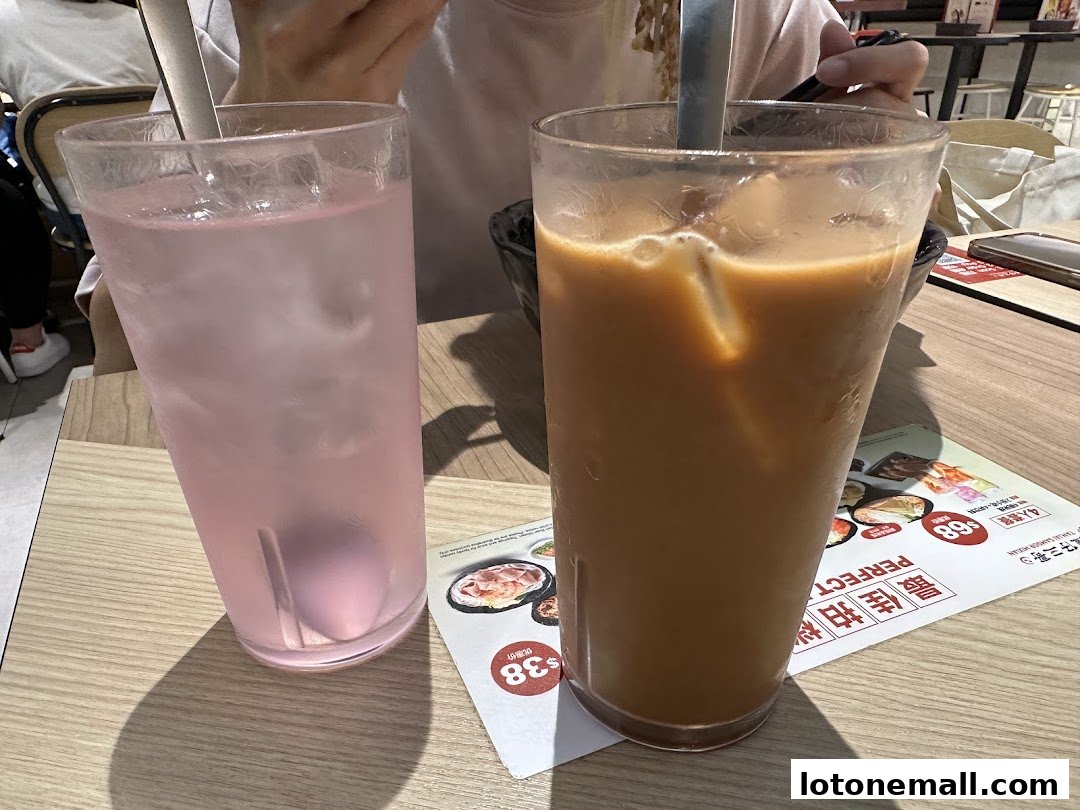 Two tall glasses of iced drinks, one pink and one milky tea, sit on a wooden table.
