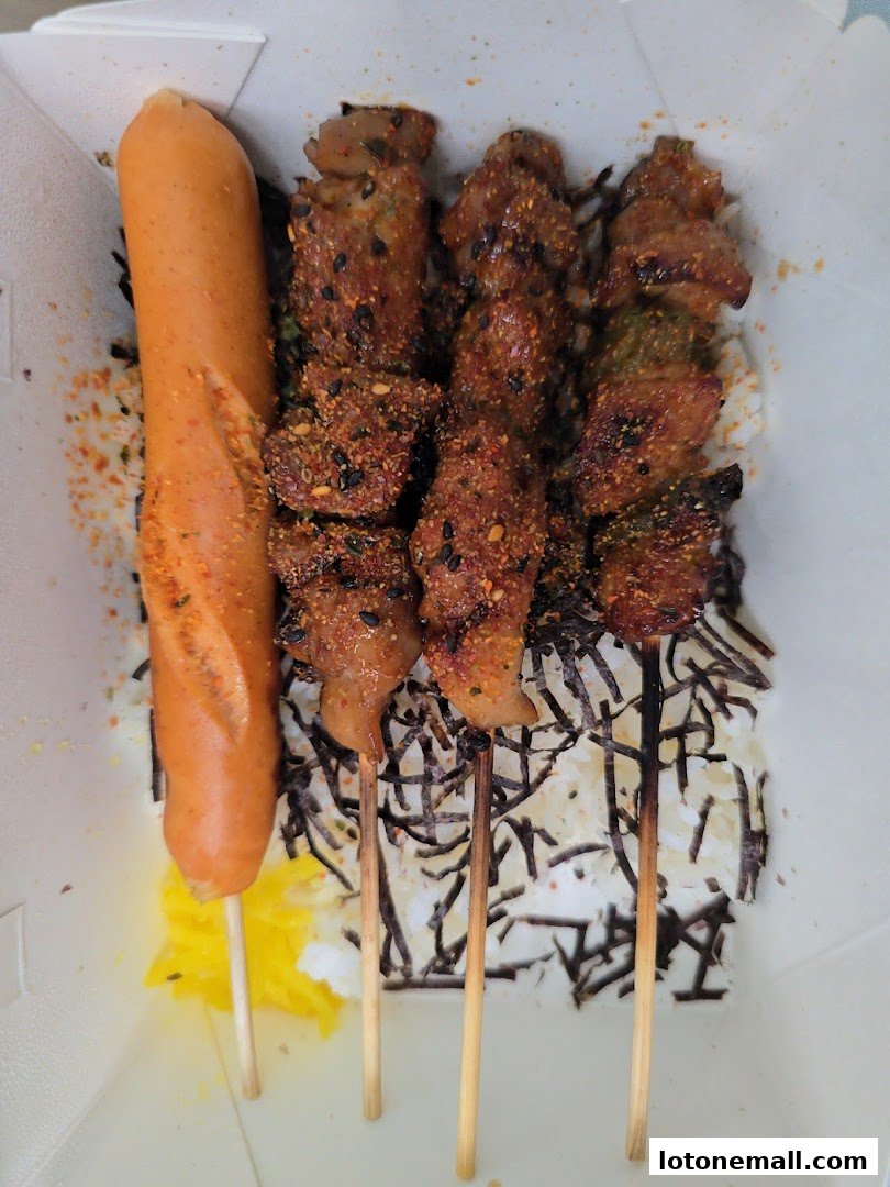 A takeout box filled with rice, seaweed, a sausage, and several skewers of seasoned meat.