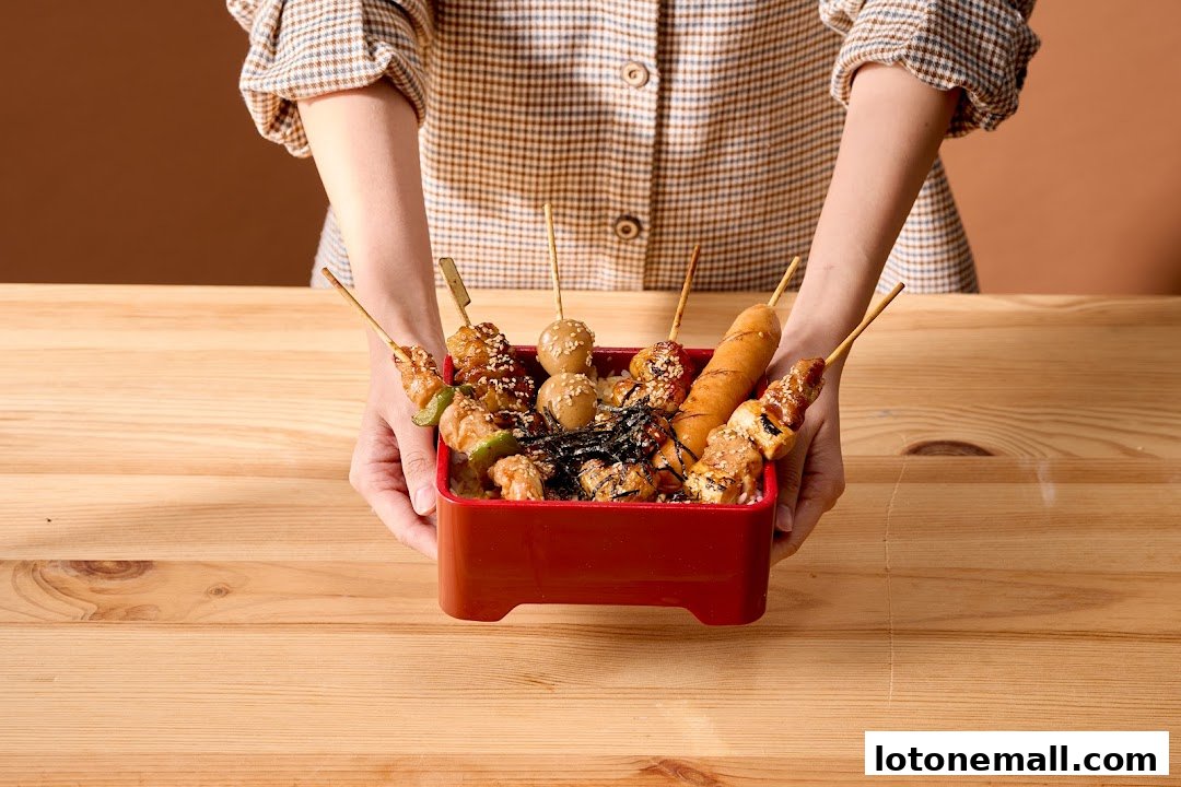 A person holds a red container filled with various yakitori skewers and rice.