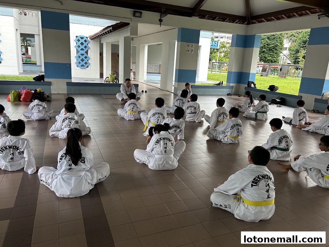 Children in Taekwondo uniforms sit on the floor during a training session.