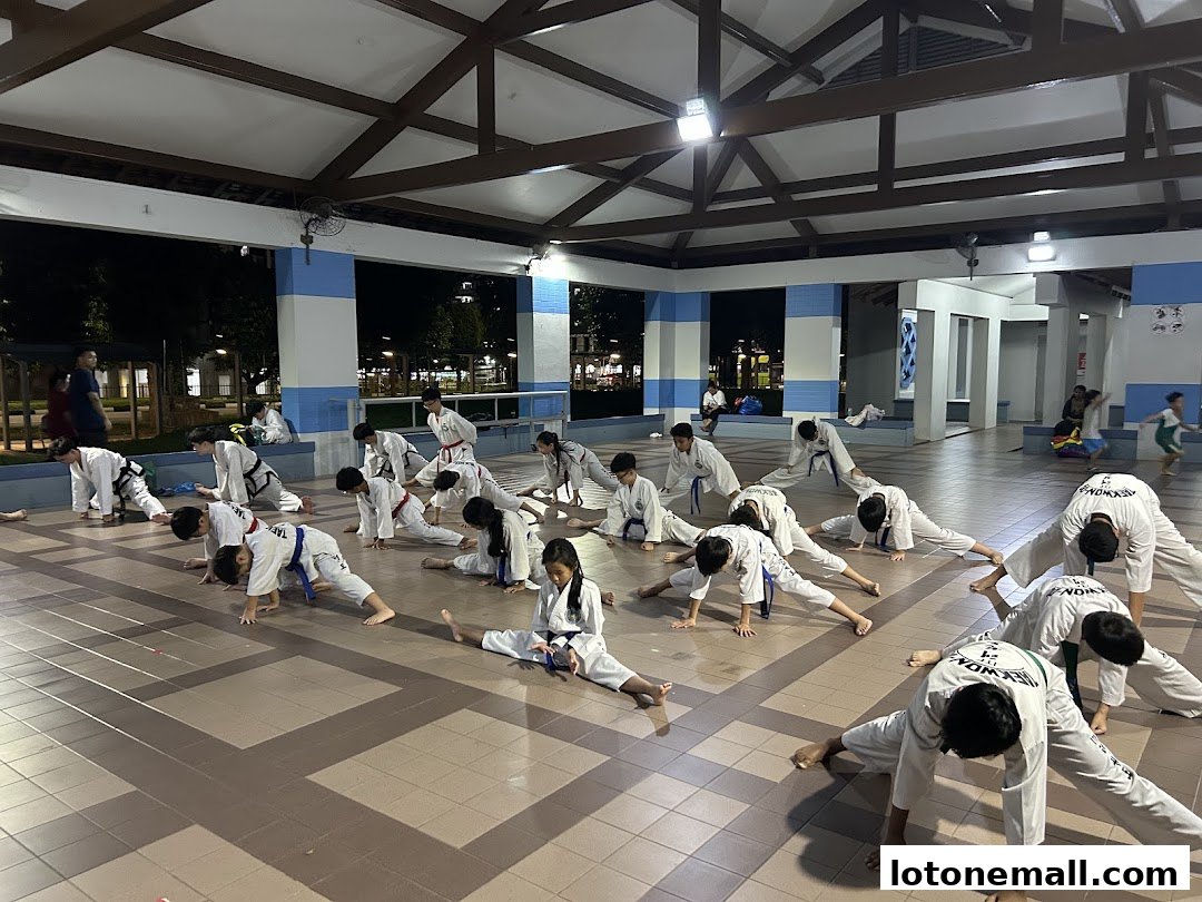 A group of children and adults in white uniforms practicing Taekwondo stretches outdoors.
