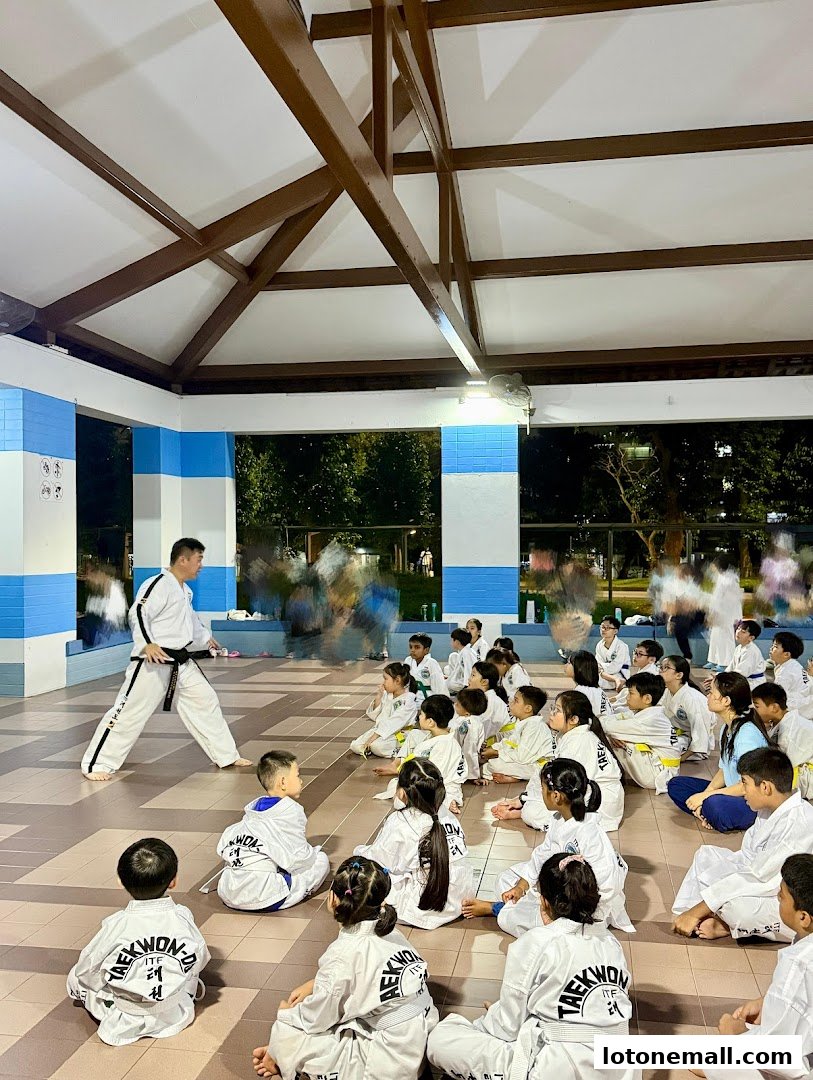 A Taekwondo instructor teaches a class of children in a covered outdoor area.