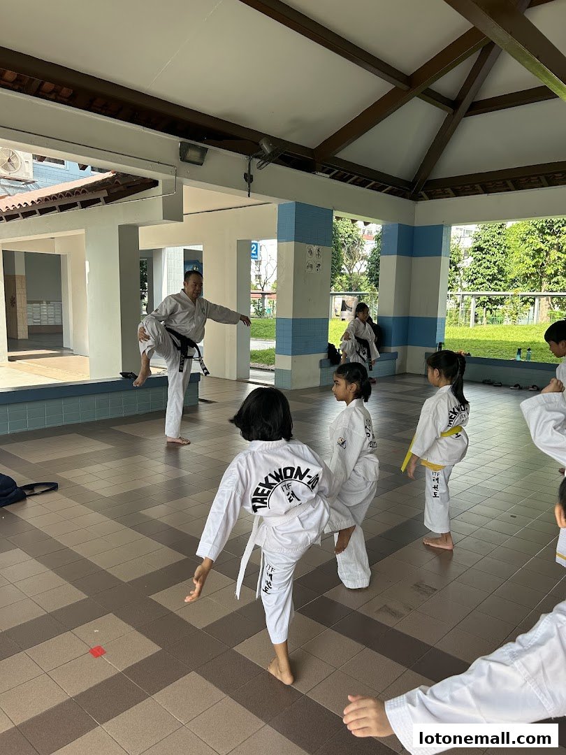 A Taekwondo instructor demonstrates a kick to students in a covered outdoor training area.