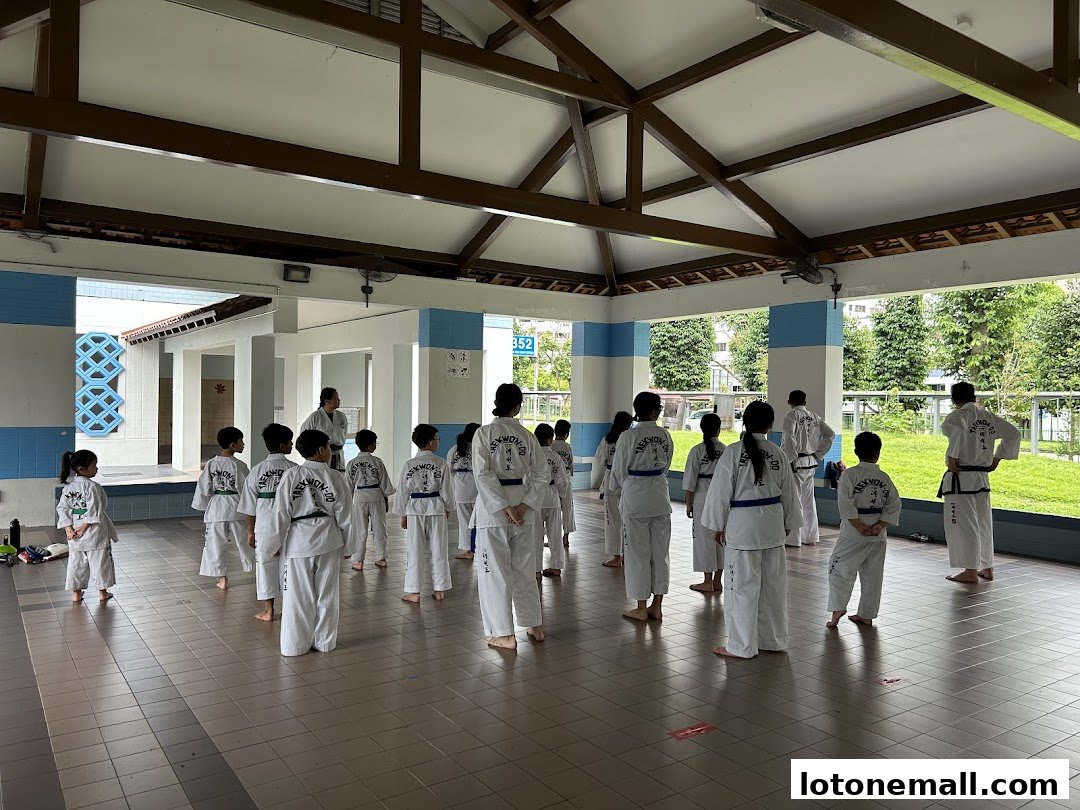 Children and adults in Taekwondo uniforms practice under a covered outdoor area.