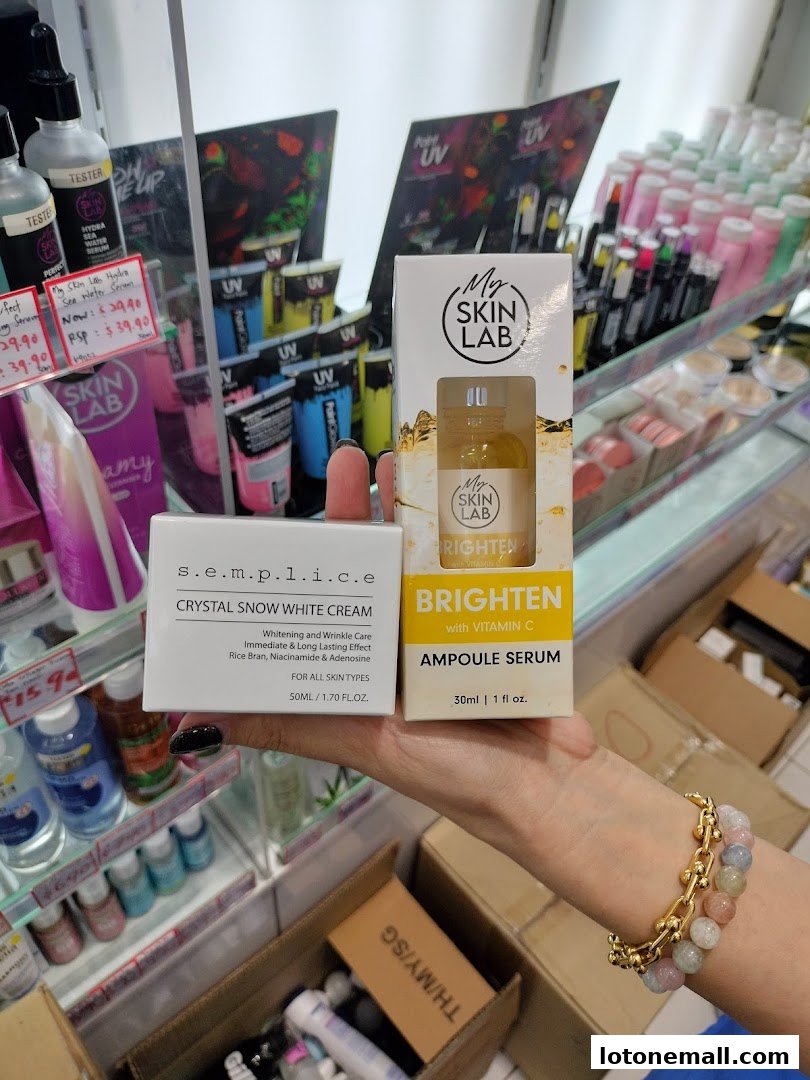 A hand holds skincare products, including a cream and serum, in front of a store display.