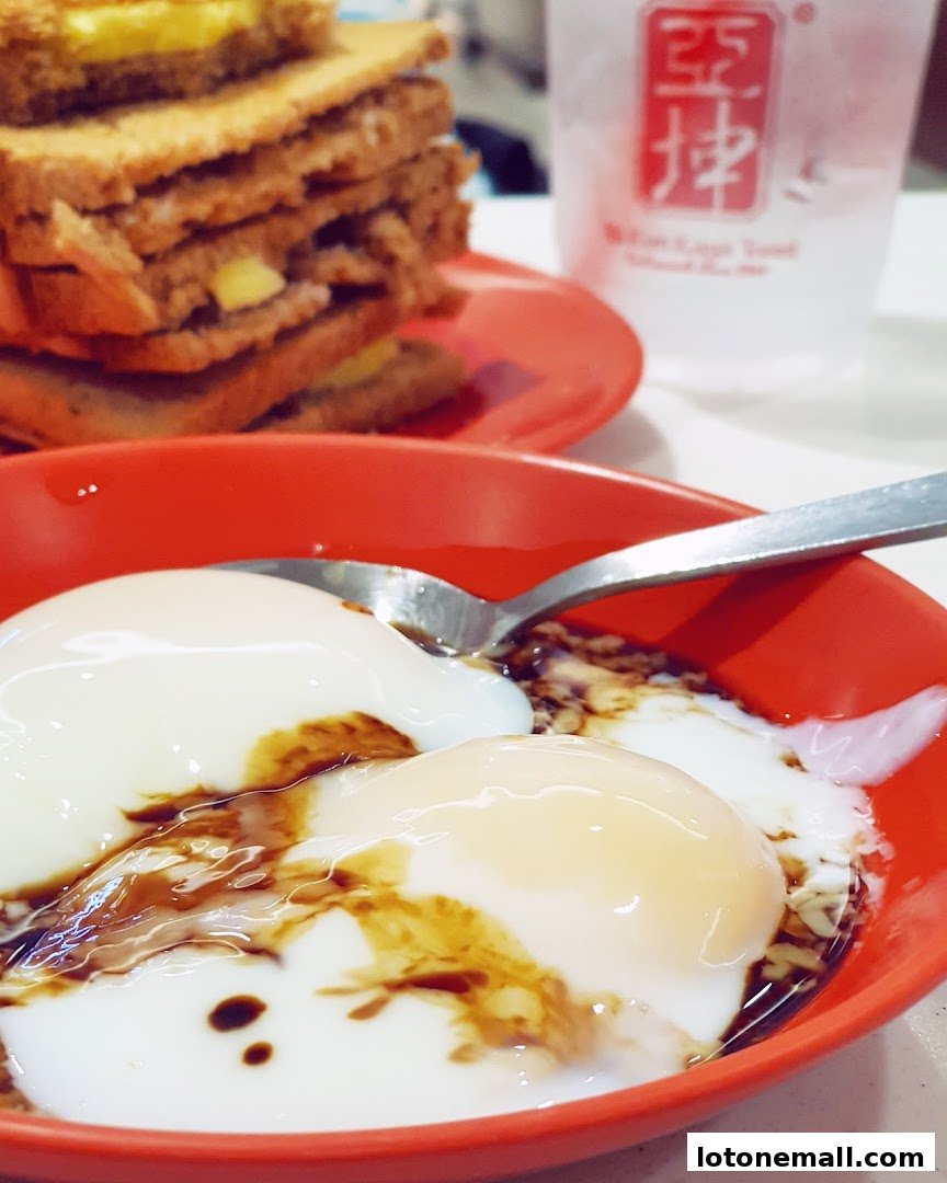 A close-up of soft-boiled eggs with soy sauce and pepper, next to kaya toast and a drink.