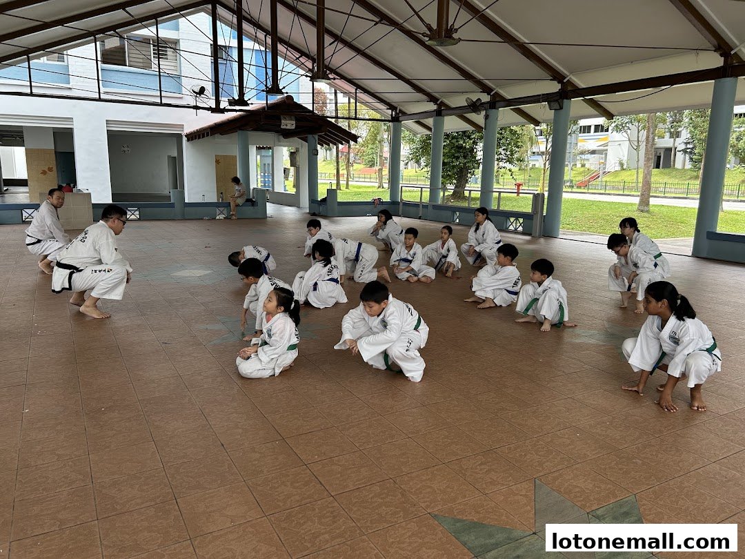 Children in Taekwondo uniforms practice with instructors in an open-air pavilion.